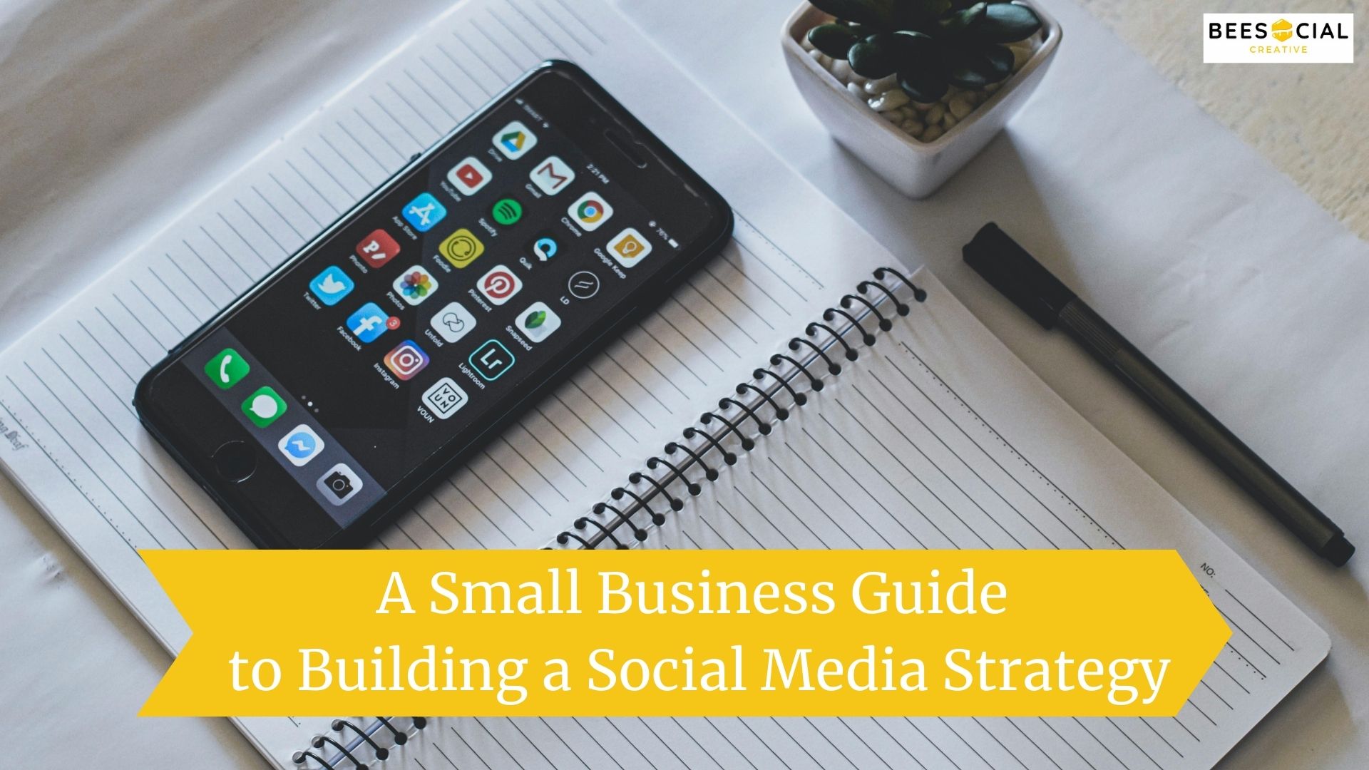 A Small Business Guide to Building a Social Media Strategy A smartphone displaying social media apps placed on an open notebook with a pen and a small plant.